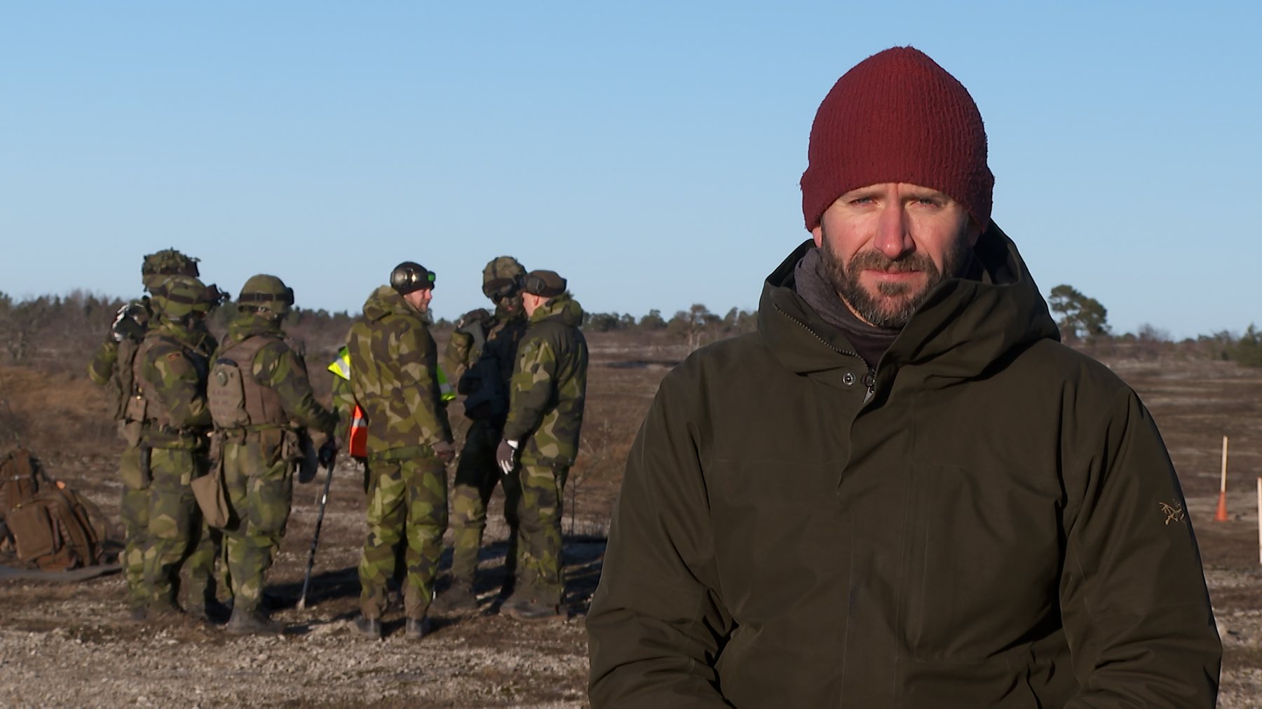 Paul Rhys reporting to camera on Gotland, Sweden, with conscripts training behind him